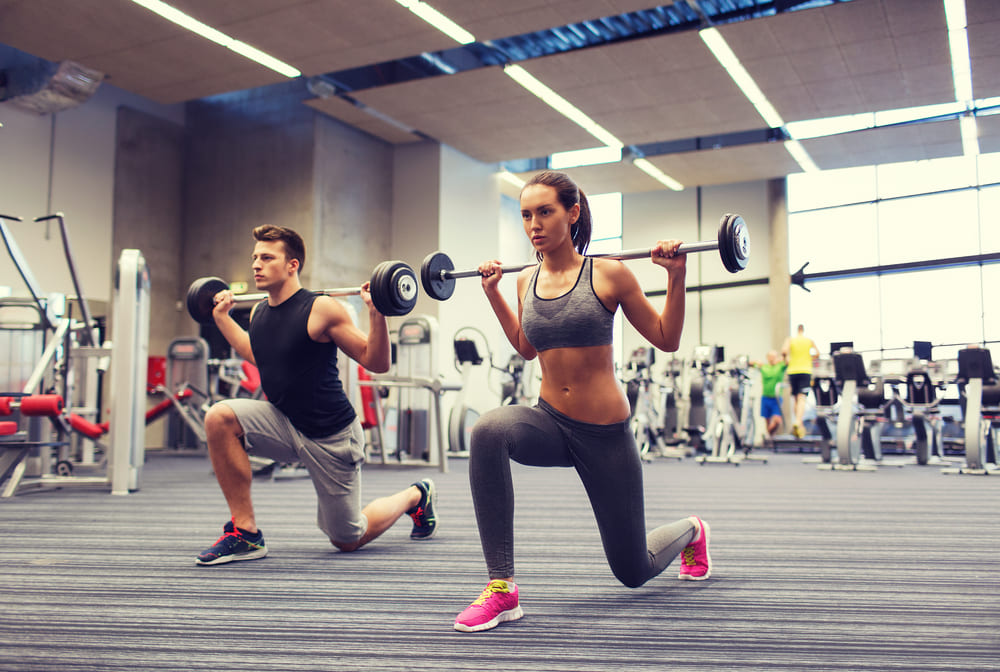 young man and woman with barbell flexing muscles and making shoulder press lunge in gym
